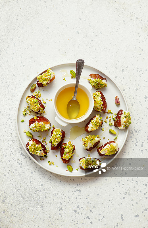 白盘子盛放着上面撒有开心果的奶油奶酪酿红枣，并淋有蜂蜜，俯视角度拍摄图片素材