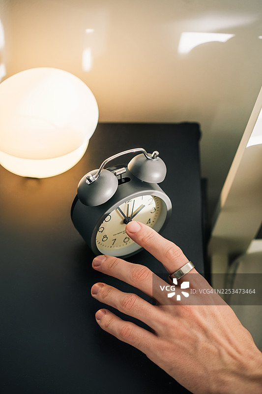 一只戴着智能戒指的手伸向床头柜上一个经典的闹钟，旁边还有一个发光的台灯。图片素材