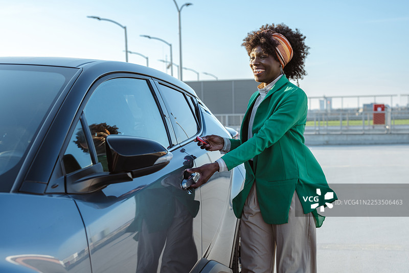 白天，一位身穿绿色西装外套的快乐女子在明亮的户外停车场解锁车门。图片素材
