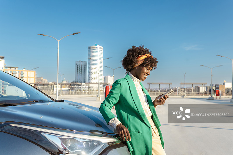 一位穿着绿色西装外套的职业女性，在阳光明媚的都市环境中倚靠在汽车旁，正在查看手机。图片素材