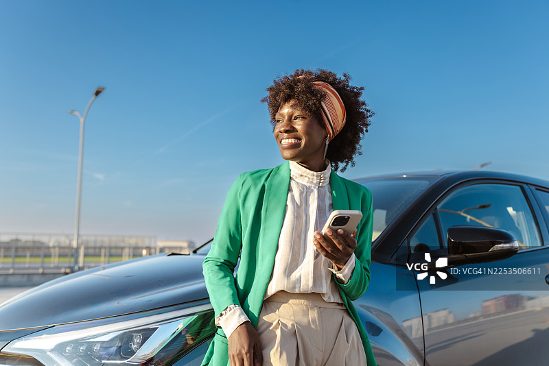 一位身穿绿色西装外套的职业女性在户外，一边使用智能手机一边微笑着站在汽车旁边。图片素材