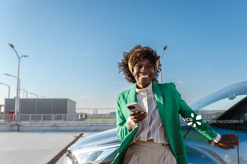 一位自信的女商人在阳光明媚的户外，身穿绿色西装外套，在汽车旁边使用智能手机。图片素材