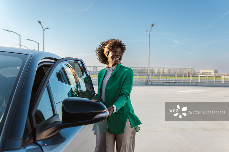 一位穿着绿色西装外套的快乐女士站在车旁，在户外享受片刻时光图片素材