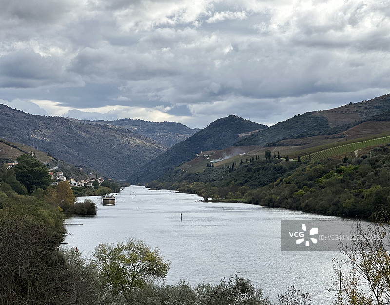 葡萄牙，在戏剧性的多云天空下，一条宽阔的河流流过山谷，山谷周围环绕着覆盖着梯田葡萄园的山丘。图片素材