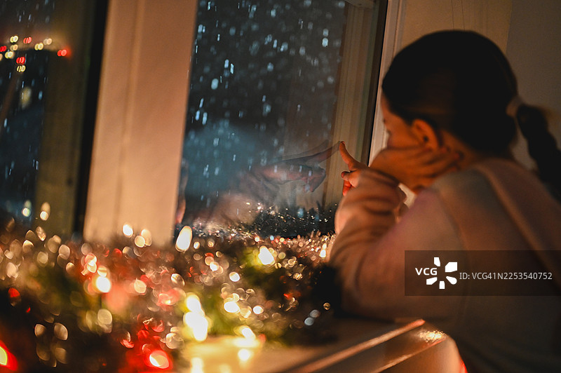 夜晚，孩子透过节日装饰的窗户看着飘落的雪花图片素材
