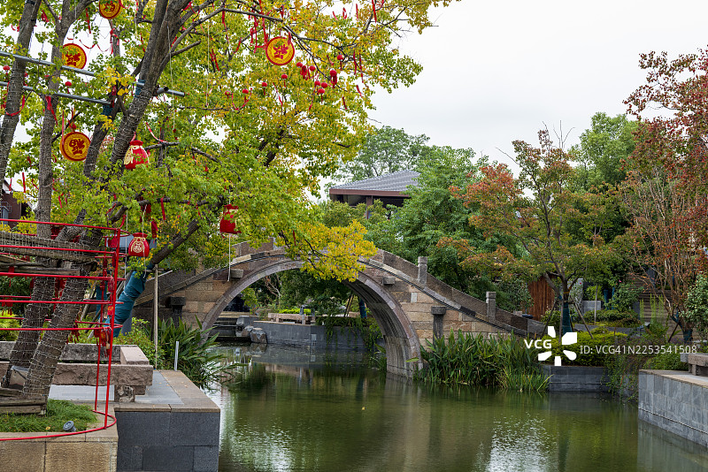 中国一座华丽的石头拱桥，跨越平静的运河，周围环绕着郁郁葱葱的绿色和红叶树木，桥的身影倒映在水中。图片素材