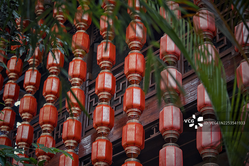 一排排传统红色中国灯笼，部分被绿色树叶遮挡，背景是木制建筑图片素材