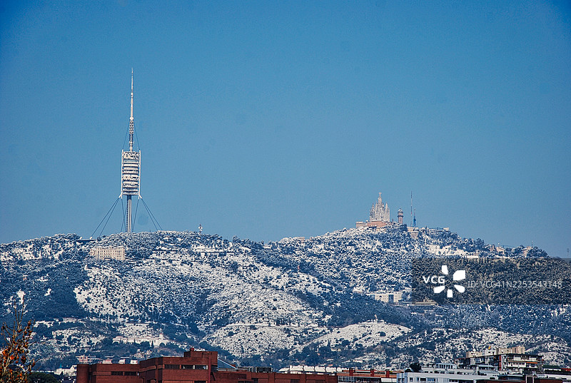 西班牙巴塞罗那，白雪覆盖的山丘上矗立着通讯塔和一座教堂，天空湛蓝图片素材