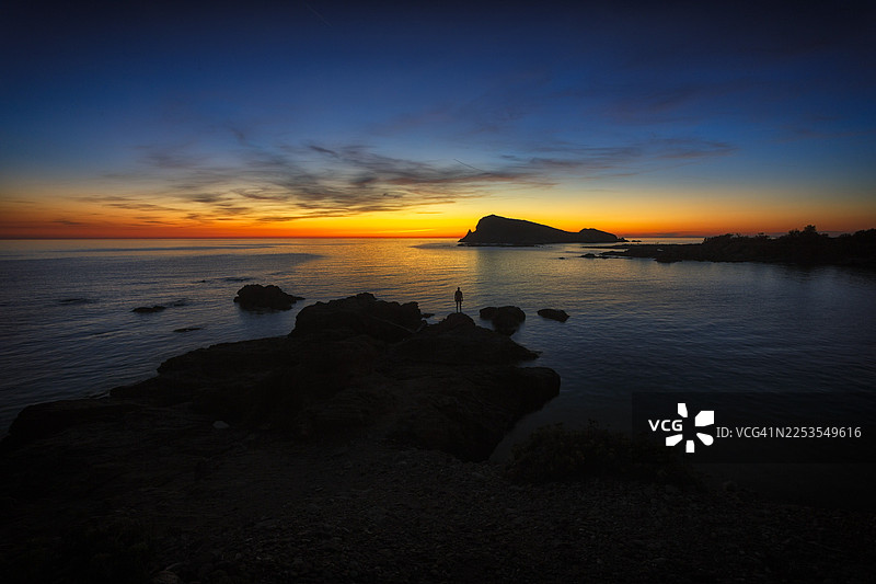 法国诺尔扎日落时分的海岸风光全景，前景为深色岩石海岸线，平静的水面倒映着充满活力的橙蓝色天空。图片素材