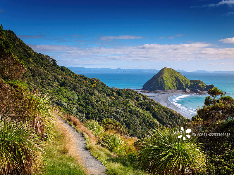 新西兰风景如画的海岸，一条蜿蜒的小路通往一个僻静的海滩和岛屿，天空湛蓝，地点为猴岛图片素材