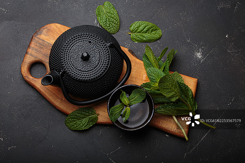 质朴木板上放着一个传统的黑色铸铁茶壶，旁边有一杯新鲜的薄荷茶和散落的薄荷叶图片素材