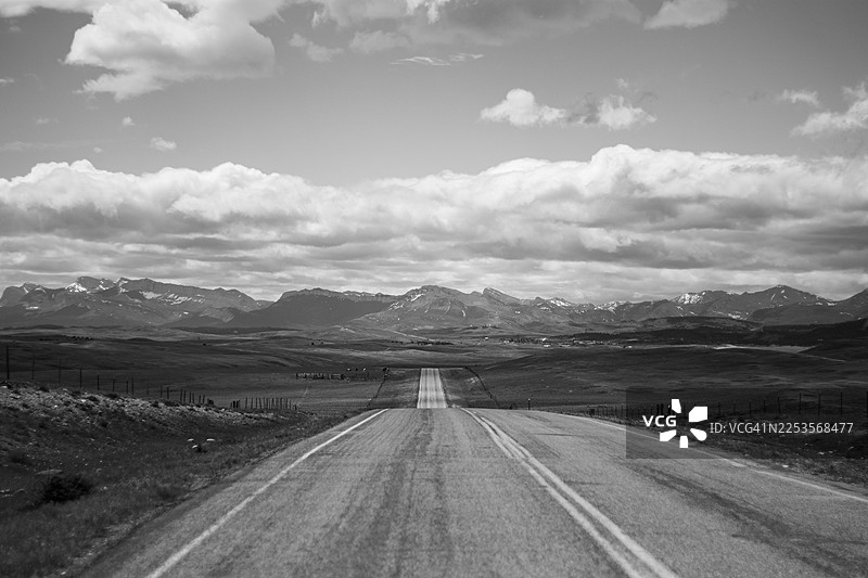 在美国，一望无际的乡村景色中，一条空旷的道路延伸至远方的山脉，天空布满云彩。图片素材