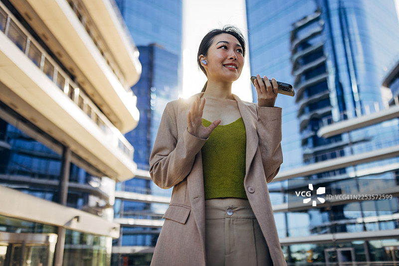 一位亚洲女商人在户外，从腰部向上拍摄，她戴着无线耳机，正用智能手机录制语音留言。图片素材