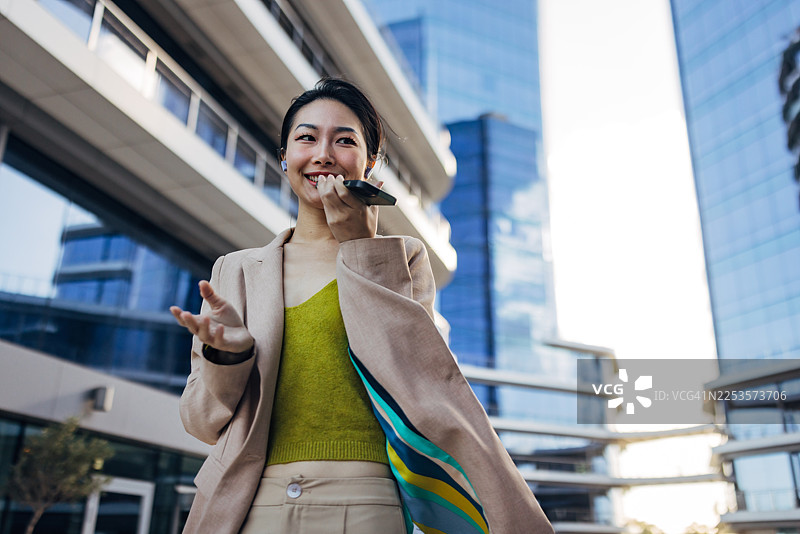 现代办公室里，一位身着商务装、面带微笑的亚洲女性，从腰部向上拍摄，她正戴着无线耳机用手机录制语音留言。图片素材