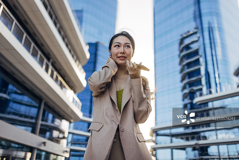 一位专业的亚洲女性在户外，戴着无线耳机，使用她的智能手机发送语音消息。图片素材