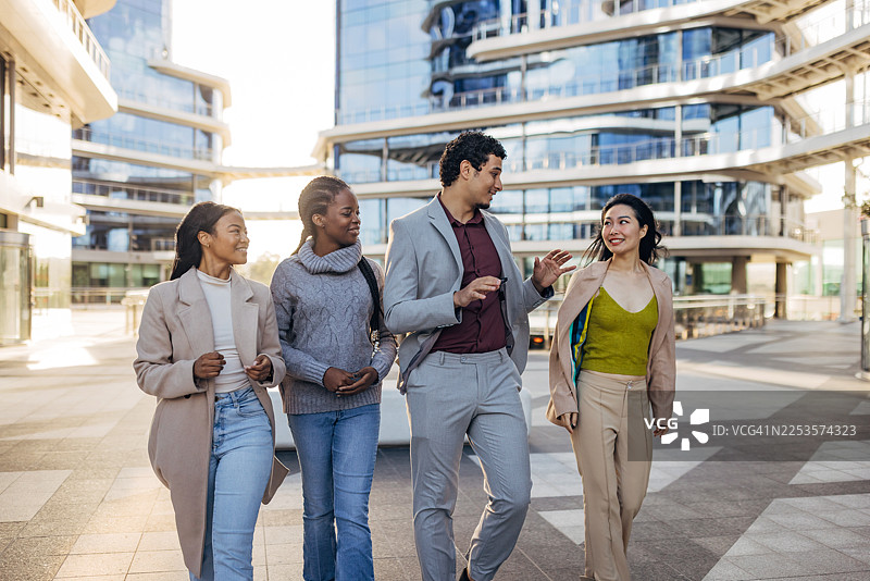 多元文化背景的同事们在现代企业大楼外一起行走交谈，这是一张展现多元化、团队合作和积极工作环境的商业生活方式图片。图片素材