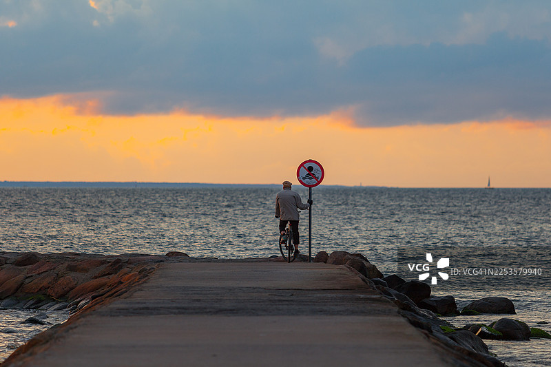 日落时分，一名男子骑着自行车经过码头，背景有禁止游泳的标志，爱沙尼亚塔林图片素材