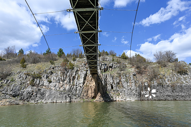 塞尔维亚，河上悬索桥的低角度视图，河岸岩石密布，有树木，蓝天背景图片素材