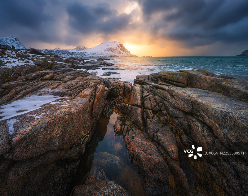 崎岖、覆雪的海岸线上，波涛汹涌的大海和乌云笼罩下的壮丽日落图片素材