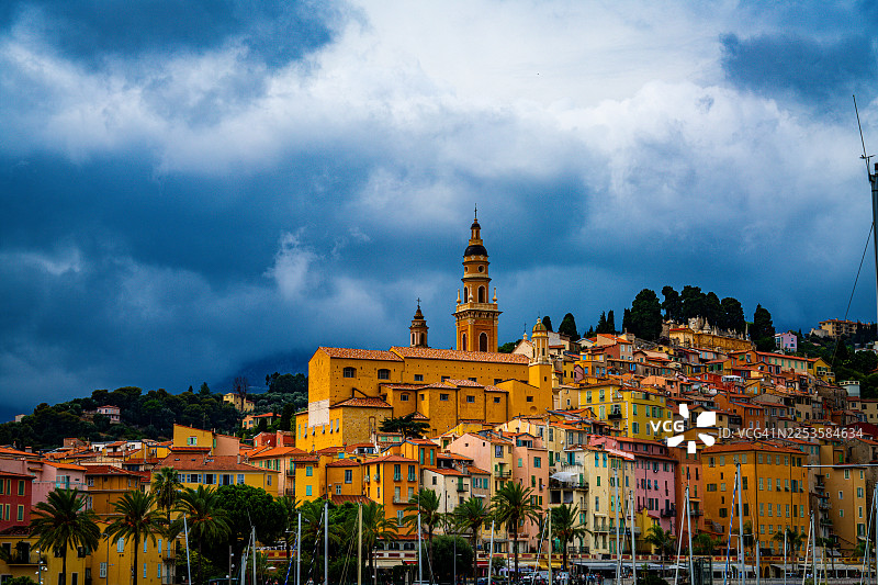法国芒通，地中海沿岸，色彩斑斓的历史建筑，一座醒目的教堂钟楼在戏剧性的多云天空下，俯瞰着地中海。图片素材