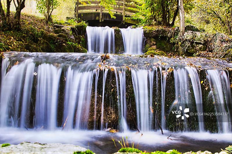 宁静的多层瀑布流经郁郁葱葱的绿色森林，背景中有一座木桥图片素材