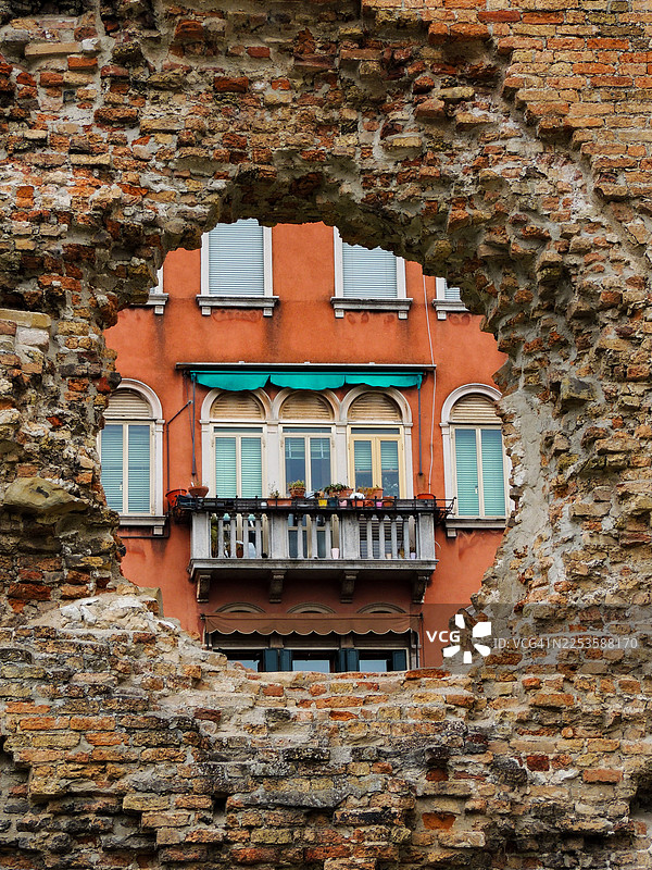 饱经风霜的砖砌拱门，框出了带有阳台和窗户的传统橙色建筑，意大利威尼斯图片素材