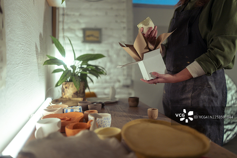 一位看不清面容的女性企业家兼陶瓷艺术家，正小心翼翼地将一个烧制完成、施釉的陶杯放入一个用牛皮纸包装的礼品盒中图片素材