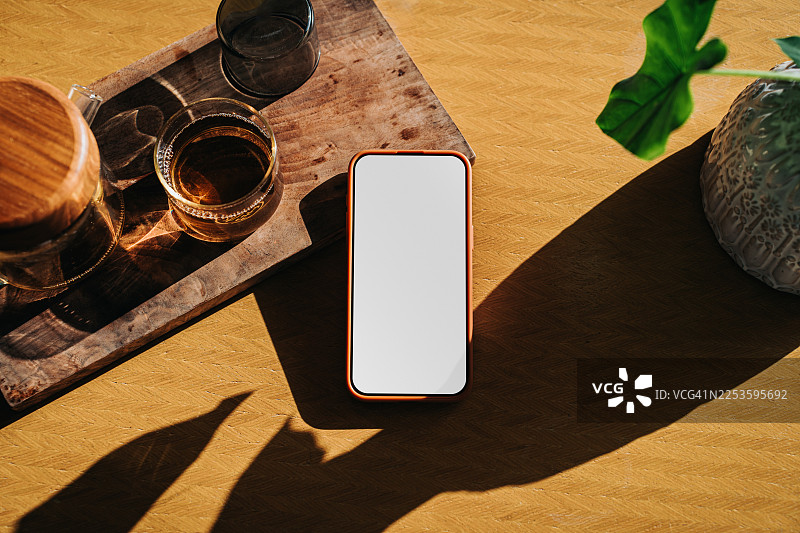 智能手机平铺在带有空白屏幕的木制办公桌上，背景是家中的阳光，旁边放着一个茶壶和一杯茶。舒适的家庭办公室工作空间，配有科技产品。手机屏幕样机图片。生活方式与科技图片素材