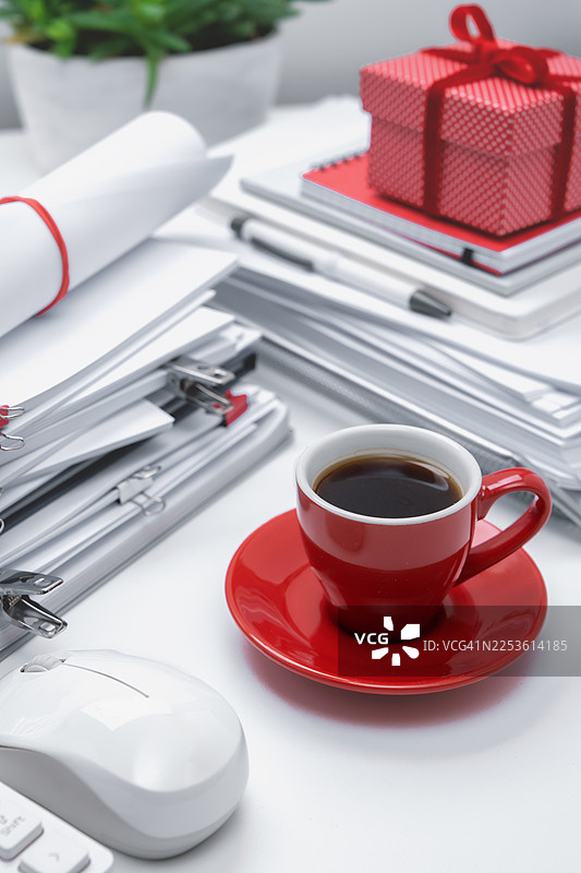 桌上的白色办公文具上放着一个红色的咖啡杯。充满纸张的办公室氛围，办公室核心。红色礼品盒图片素材