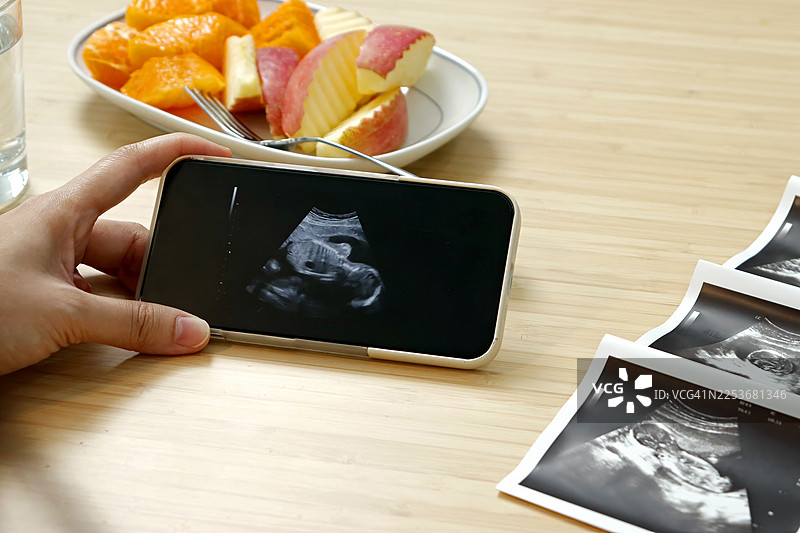 孕妇在手机上查看胎儿超声波扫描图，旁边放着一盘新鲜切片水果，象征健康孕期饮食图片素材