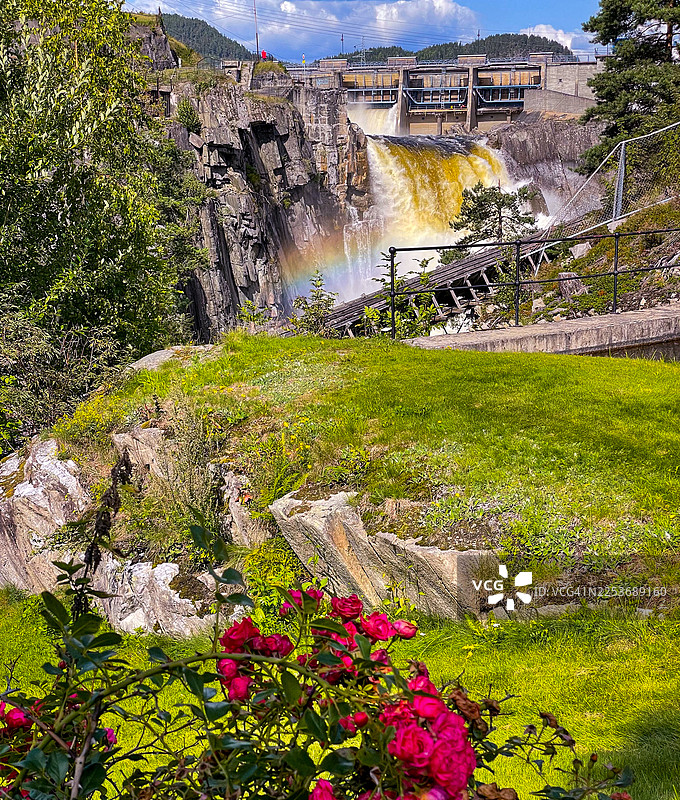 前景是模糊的红色玫瑰和绿色草坪的景观。背景是水坝和发电站的瀑布。部分旧木制水道可见。图片素材