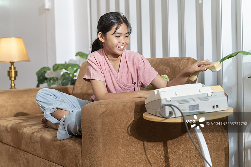 快乐的亚洲十来岁女孩在家里的棕色沙发上，通过固定电话放松地交谈，用于沟通概念图片素材