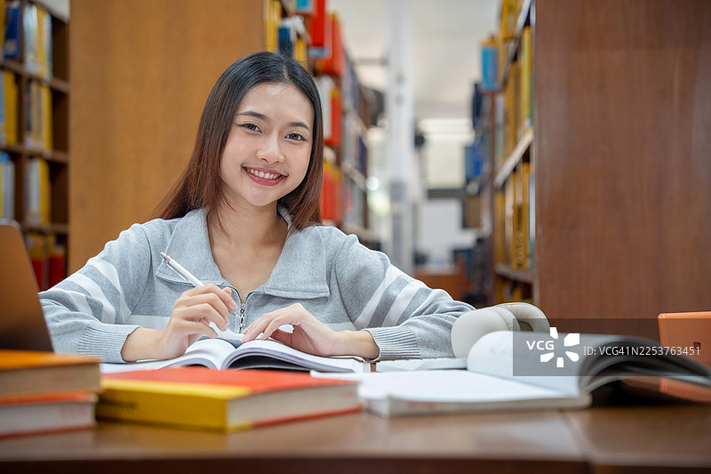 教育与学校概念。年轻女子在图书馆读书。一位漂亮的亚洲女大学生微笑着，手里拿着书，坐在图书馆里。学习与教育。图片素材