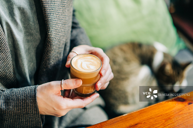 一位身穿时尚灰色西装外套的人，手持一杯拿铁拉花咖啡，背景中有一只熟睡的猫。图片素材