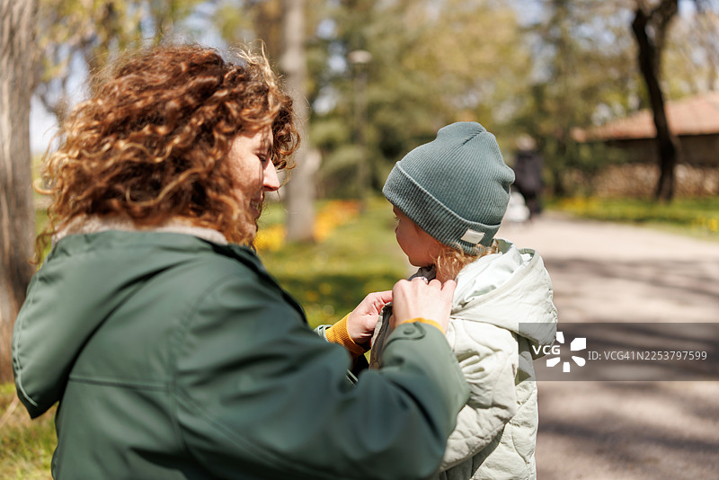 在一个寒冷的初春公园里，一位慈爱的母亲正在为女儿扣上外套的纽扣图片素材