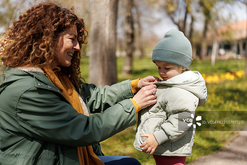 在一个寒冷的初春，一位母亲在公园里为女儿扣上外套的纽扣图片素材