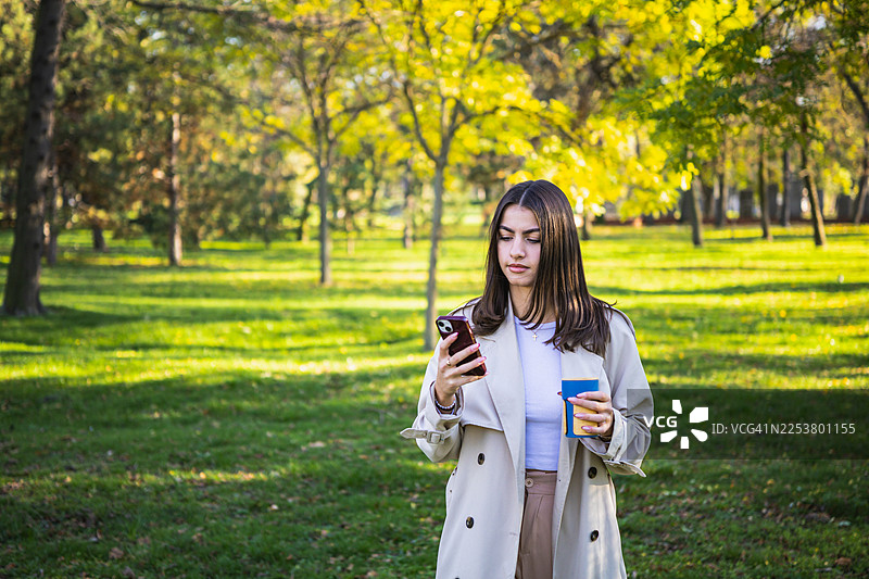 秋日暖阳下，一位身穿外套的美丽年轻女子在公园的咖啡休息时间使用手机。图片素材