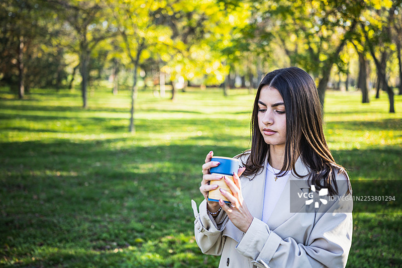 秋日暖阳下，一位身穿外套的年轻美丽女子在公园的咖啡休息时间肖像。图片素材