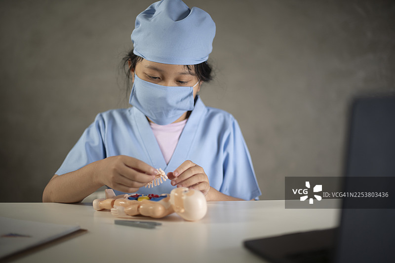 一个穿着医疗服、戴着口罩的孩子正在仔细地检查一个玩偶，仿佛一位医护人员在练习医疗操作。图片素材