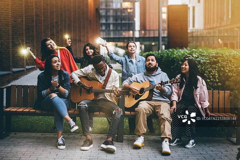 一群充满活力的不同肤色的朋友在黄昏时分聚集在一张木制长椅上，弹奏着原声吉他放声歌唱。温暖的城市灯光和快乐的表情捕捉到了都市社区和共同音乐热情的精髓。图片素材