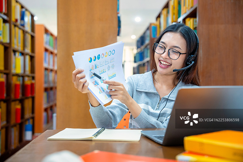 一名亚洲女学生在图书馆通过电脑笔记本进行视频会议，与老师进行在线学习。返校季，教育、知识、大学。戴着耳机的女学生在图书馆使用笔记本电脑和平板电脑工作。学习与学校概念。图片素材