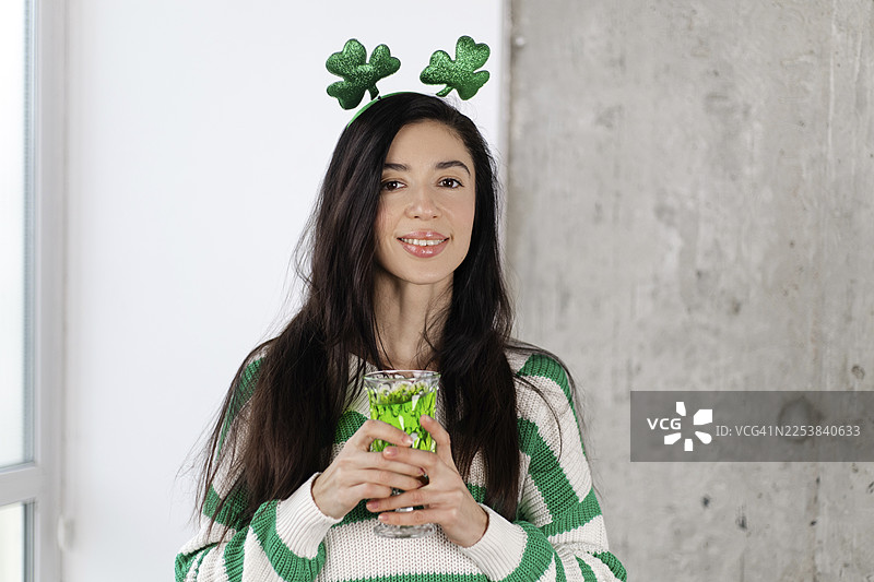一位身穿圣帕特里克节主题服装、手持盛满绿色饮品的玻璃杯的年轻女子，脸上洋溢着喜悦。图片素材