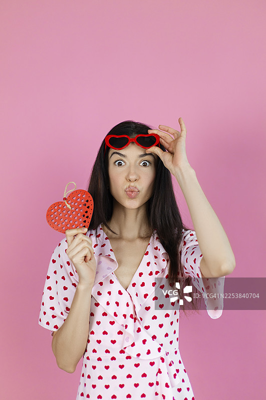 一个年轻活泼的女子站在粉色背景前，她一手拿着红心装饰，一手戴着心形太阳镜在额头上。图片素材