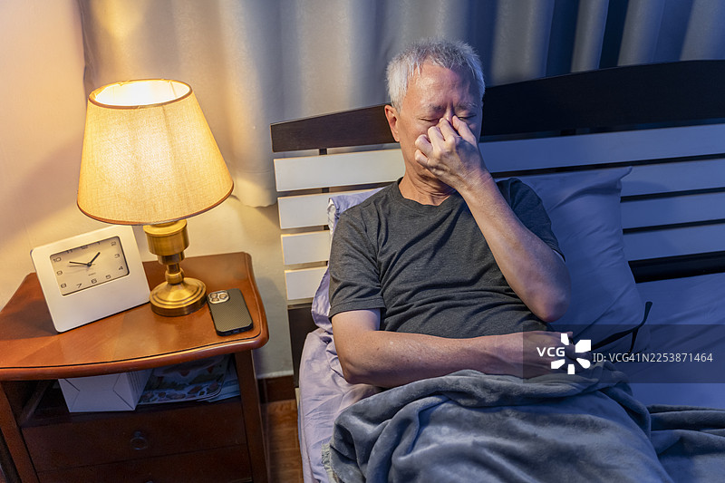 晚上，一位年长的亚洲男性在床上使用智能手机后揉着疲惫的眼睛图片素材