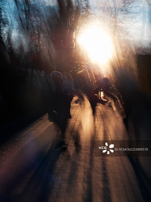日落时分，行人沿着铺砌的小路行走，画面因刻意运镜而产生动态模糊的剪影。低垂的太阳强烈的逆光效果，勾勒出长长的影子，光线如同发光的条带，营造出抽象的都市氛围。图片素材