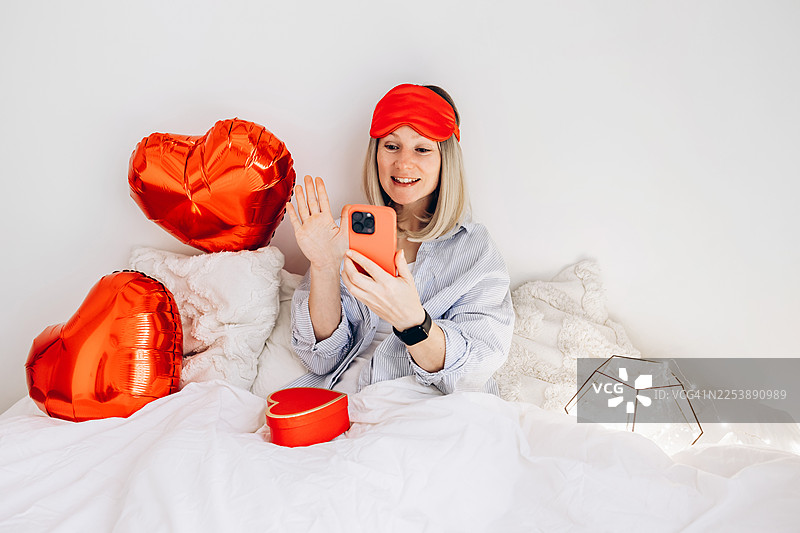 躺在床上的女人在进行智能手机视频通话时挥手，白色床上用品上有红心气球和心形礼品盒，情人节异地恋和在线问候的概念图片素材