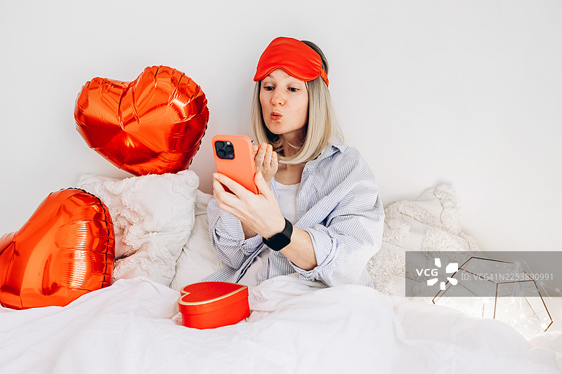 女人坐在床上，通过智能手机发送飞吻。情人节在线交流和浪漫信息概念图片素材