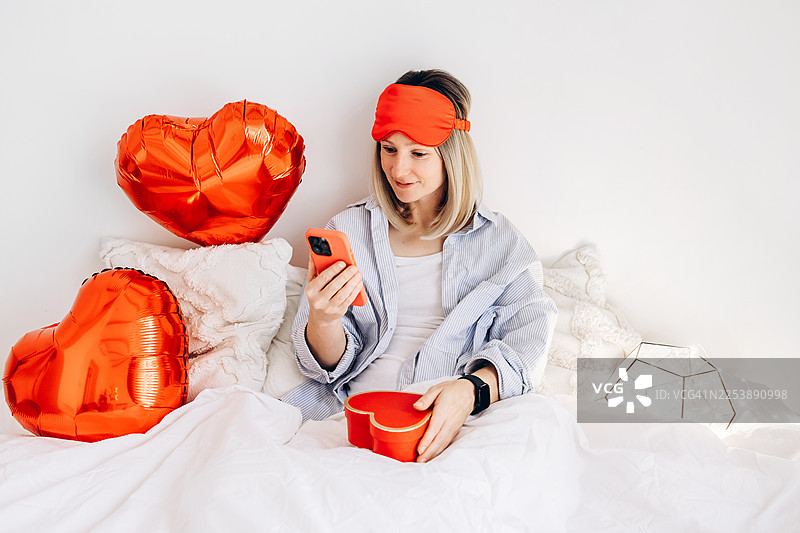 女人坐在床上，手持红色心形礼品盒和智能手机，周围环绕着心形气球和舒适的卧室装饰。图片素材