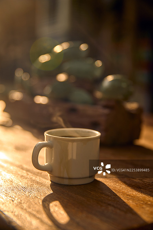 阳光照射下的木桌上放着一杯冒着热气的白色咖啡杯。图片素材