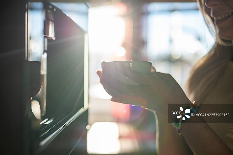 一位年轻的、不为人知的女性正在享用咖啡机制作的新鲜热咖啡，晨光透过窗户洒入，蒸汽袅袅升起，这是一个温馨的居家时刻。图片素材